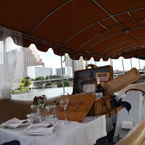 View of table with food, wine glasses and cutting board in front of picnic basket. Lake and bridge in the background from inside a small boat with orange roof