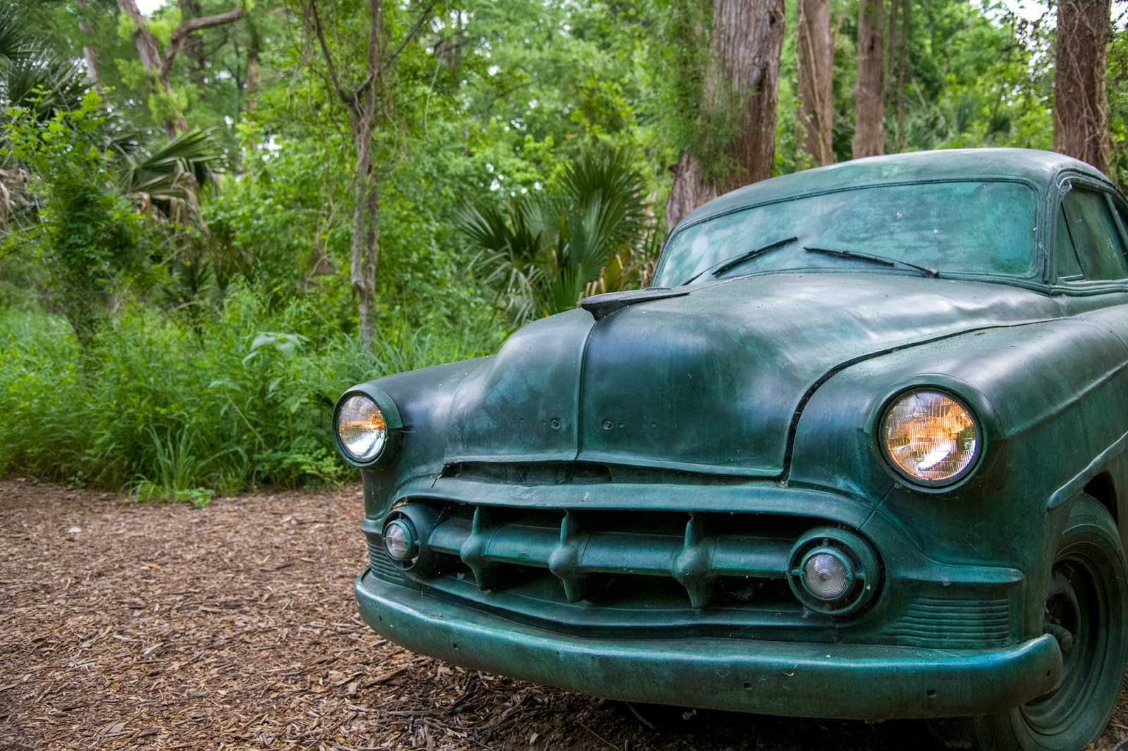 Sculpture of a retro style car that has patina. Headlights on. Green growth in the background.