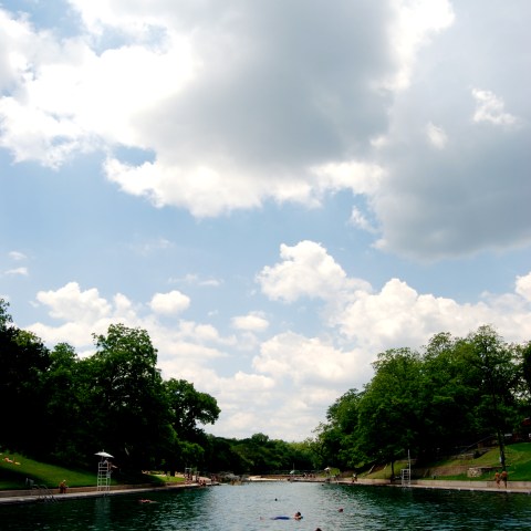 People laying on floats in Barton Springs Pool. The water is a blue-green color, green trees line the edges of the pool, and clouds are in the sky with blue sky