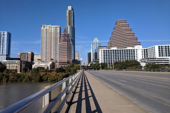 View looking down a bridge at the Texas State Capital with Downtown buildings on either side. Blue sky.