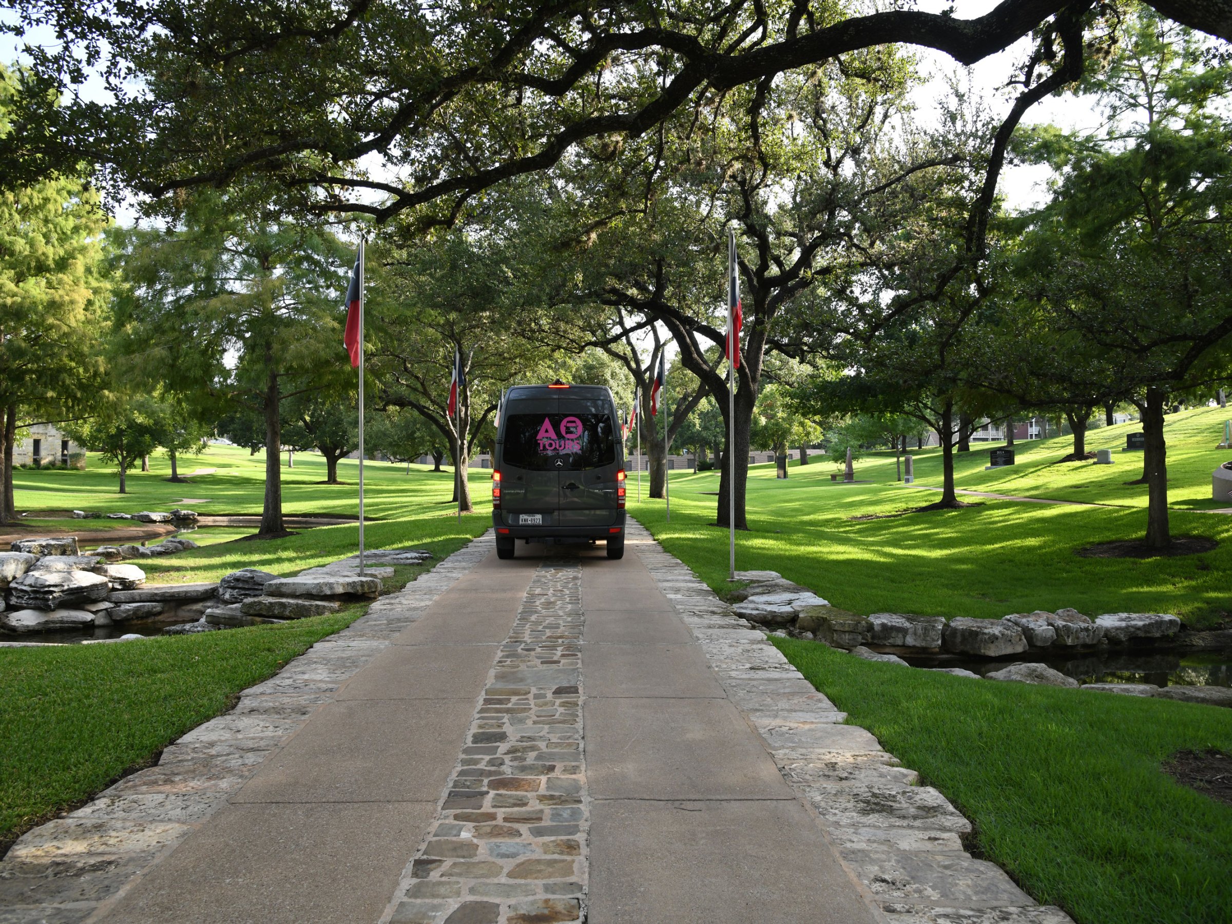 Dark Gray AO Tours Austin Sprinter Van on a small road with Texas Flags along the side