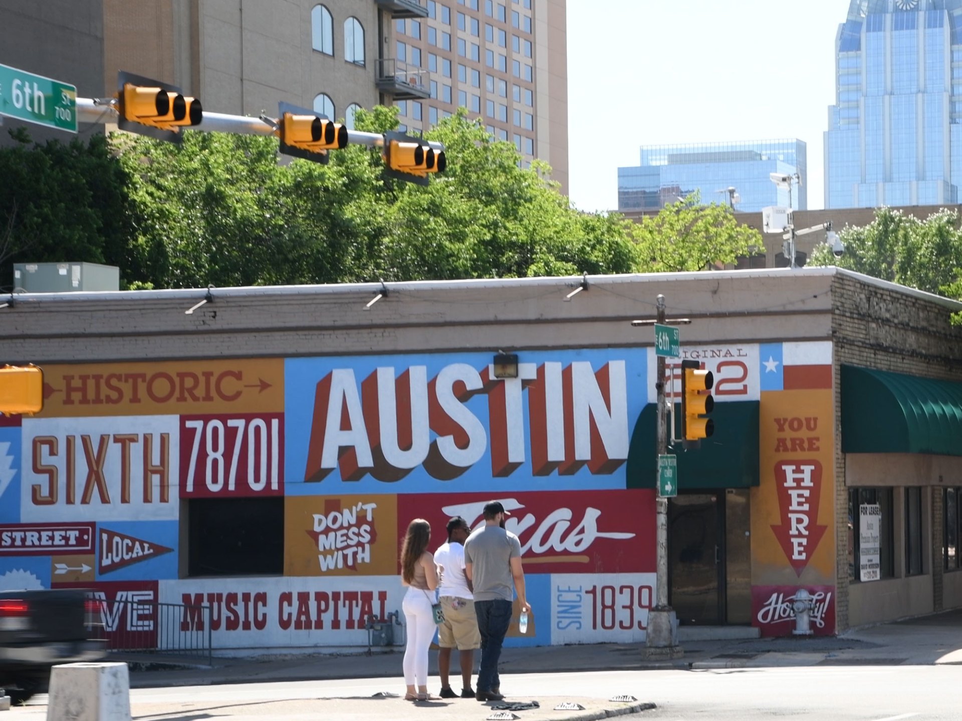 Austin painted mural on side of a building. Taller buildings in the background. People waiting to cross the street at the corner.