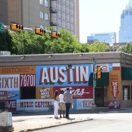 Austin painted mural on side of a building. Taller buildings in the background. People waiting to cross the street at the corner.
