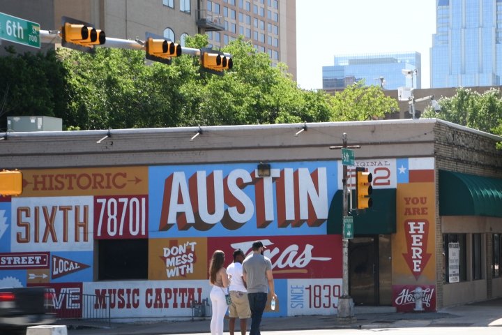 Austin painted mural on side of a building. Taller buildings in the background. People waiting to cross the street at the corner.