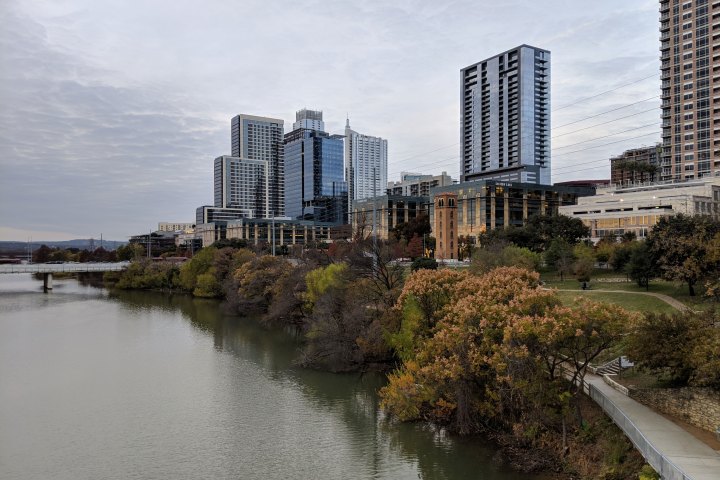 a body of water with Downtown Austin in the background. Autumn color leaves on the trees, cloudy gray sky.