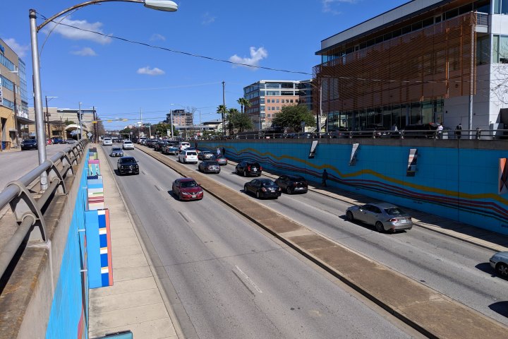 View from the bridge above a road with traffic going both directions. Painted walls on both sides of the street that are blue with yellow, red, pink and purpled pained lines. Blue sky.