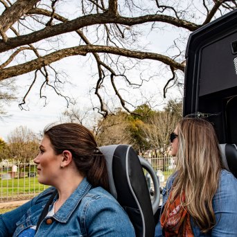 Two passengers looking out the side of the AO Tours Austin Panoramic Bus with the top down. Trees and fence in the background.
