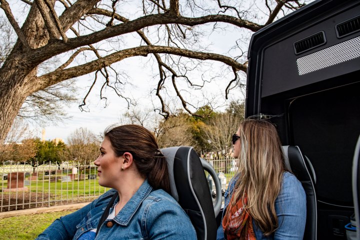 Two passengers looking out the side of the AO Tours Austin Panoramic Bus with the top down. Trees and fence in the background.