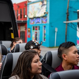 Hispanic woman and South East Asian man sitting together looking out the side of the of the bus with the top down. White woman with blonde hair and sunglasses looking the opposite direction. Blue, red, pink, white, and black painted murals on the building walls.