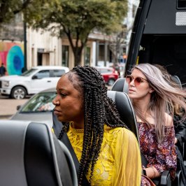 Group of people looking street murals from AO Tours Austin Panoramic Van. African American woman in front looking out the side of the open roof. Woman behind with rose colored sunglasses and blond hair blowing in the wind. Multicolor mural in the background.