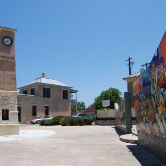 Mosaic mural made up of orange, yellow, purple, blue, brown, and white tiles. Clock tower and buildings next to it. Clear blue sky.