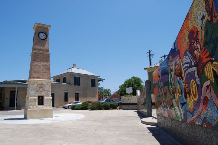 Mosaic mural made up of orange, yellow, purple, blue, brown, and white tiles. Clock tower and buildings next to it. Clear blue sky.