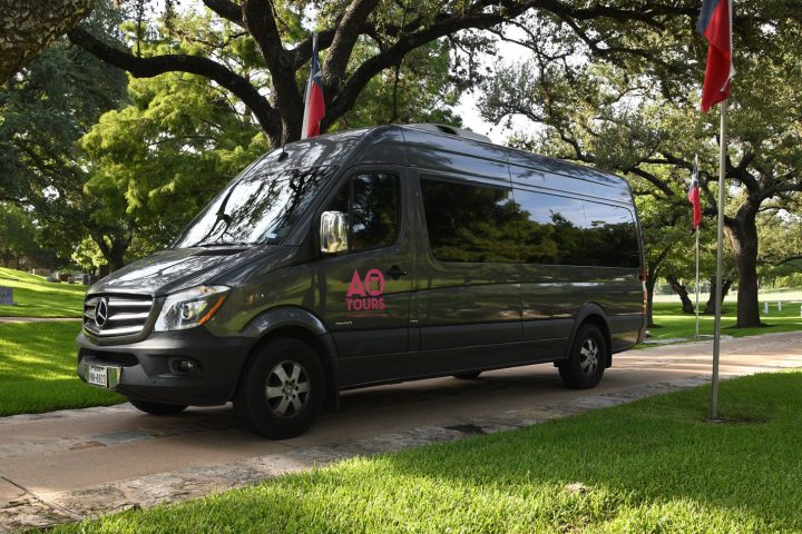 Dark Gray AO Tours Austin Sprinter Van on a small road with Texas Flags along the side