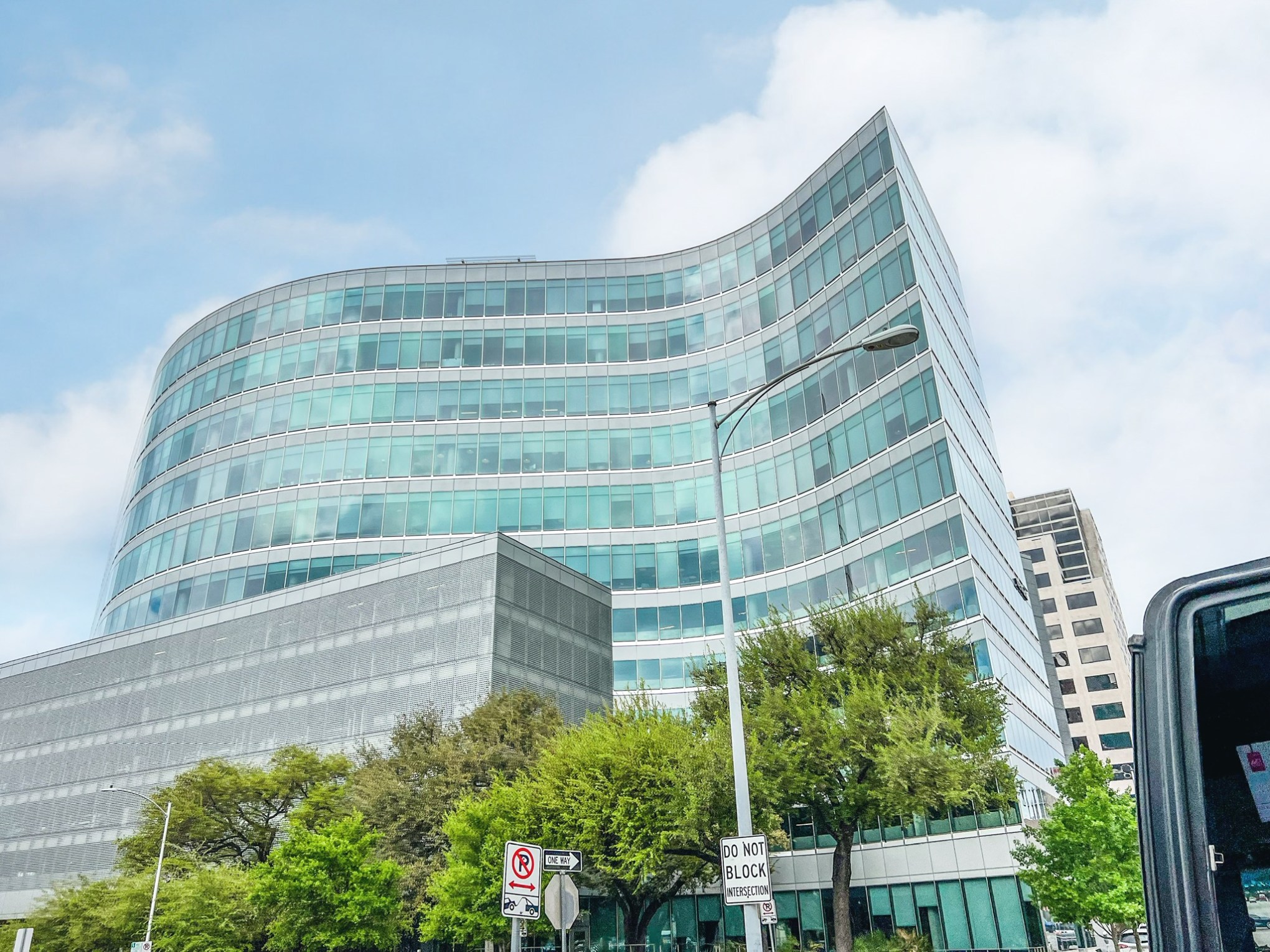 A tall glass building in a Austin, Tx. Trees growing up in front. Blue sky with white clouds.