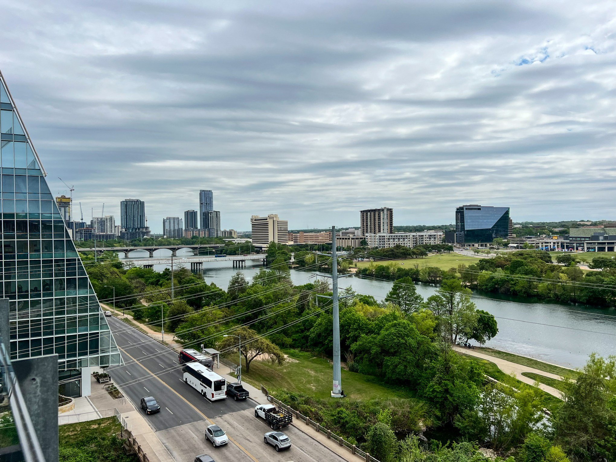 Congress and 1st bridges going over a Ladybird Lake in Austin, TX. Cloudy overcast sky, green trees, cars and a bus on the street. The corner of a tall glass building. Tall buildings in the distance.
