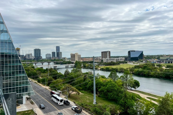 Congress and 1st bridges going over a Ladybird Lake in Austin, TX. Cloudy overcast sky, green trees, cars and a bus on the street. The corner of a tall glass building. Tall buildings in the distance.