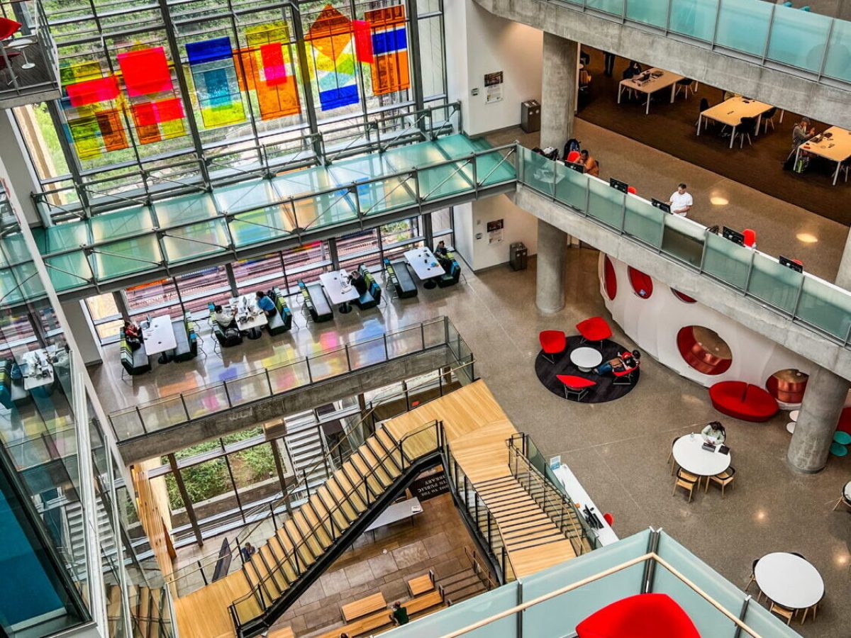 View from top floor of the Austin Central Public Library looking down through the central atrium. Red chairs, white tables, wood stairs, glass windows, multicolor abstract art hanging in the space.