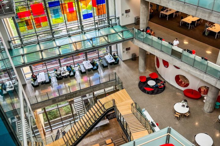 View from top floor of the Austin Central Public Library looking down through the central atrium. Red chairs, white tables, wood stairs, glass windows, multicolor abstract art hanging in the space.