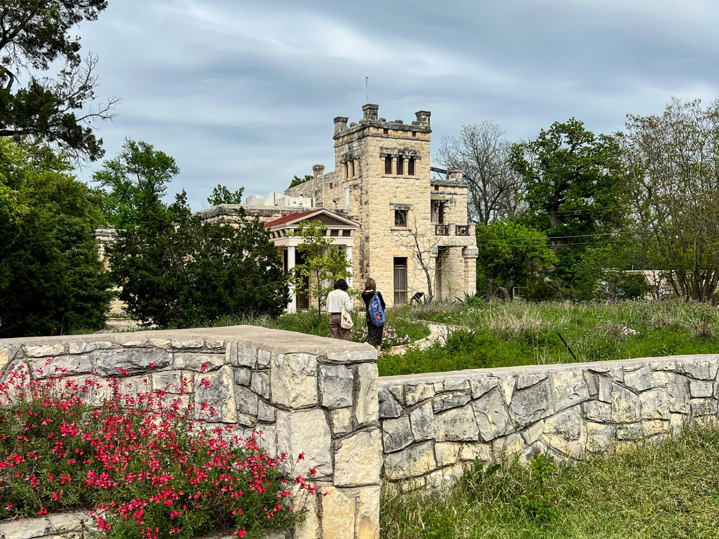 View of the Elisabet Ney Museum. Old light stone wall in the foreground with pink flowers and grass growing. Castle like historic building in the background. Cloudy overcast sky.