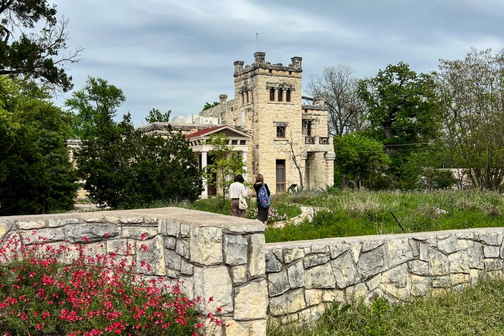 View of the Elisabet Ney Museum. Old light stone wall in the foreground with pink flowers and grass growing. Castle like historic building in the background. Cloudy overcast sky.