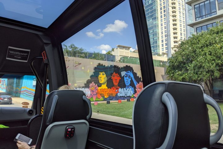 View from inside AO Tours Austin Panoramic Bus. Mural of three different woman on a wall, one yellow, one red, and one blue. Blue sky and green trees and grass around.